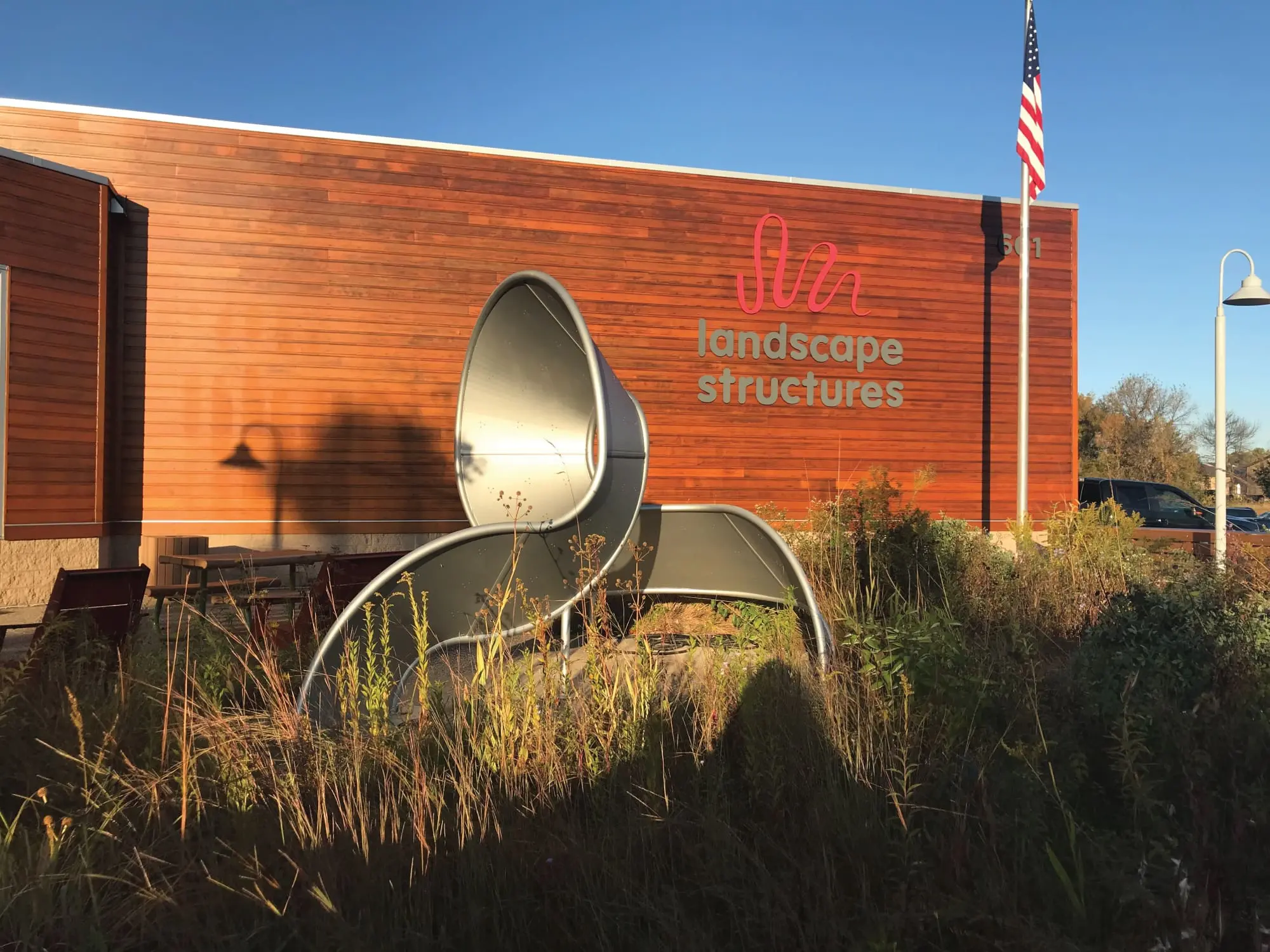 Modern metal sculpture outside a building with landscape structures sign, American flag, and clear blue sky.