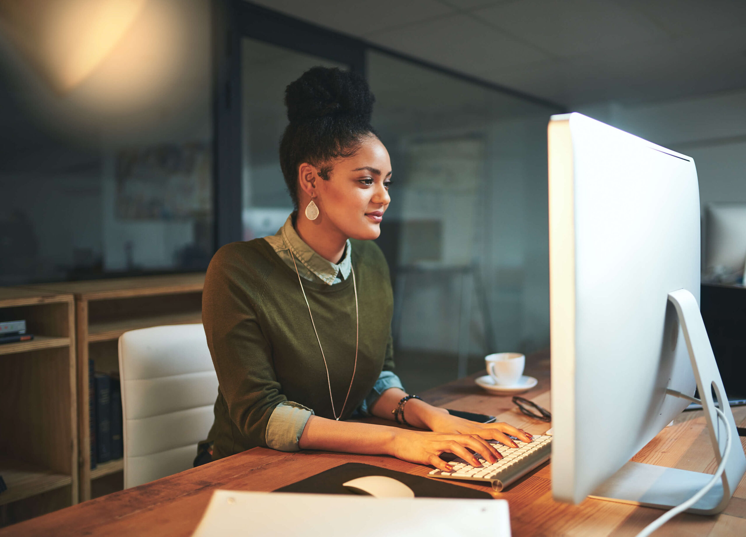 Woman working at a computer in a cozy office setting, focusing on a task, with a coffee cup nearby.