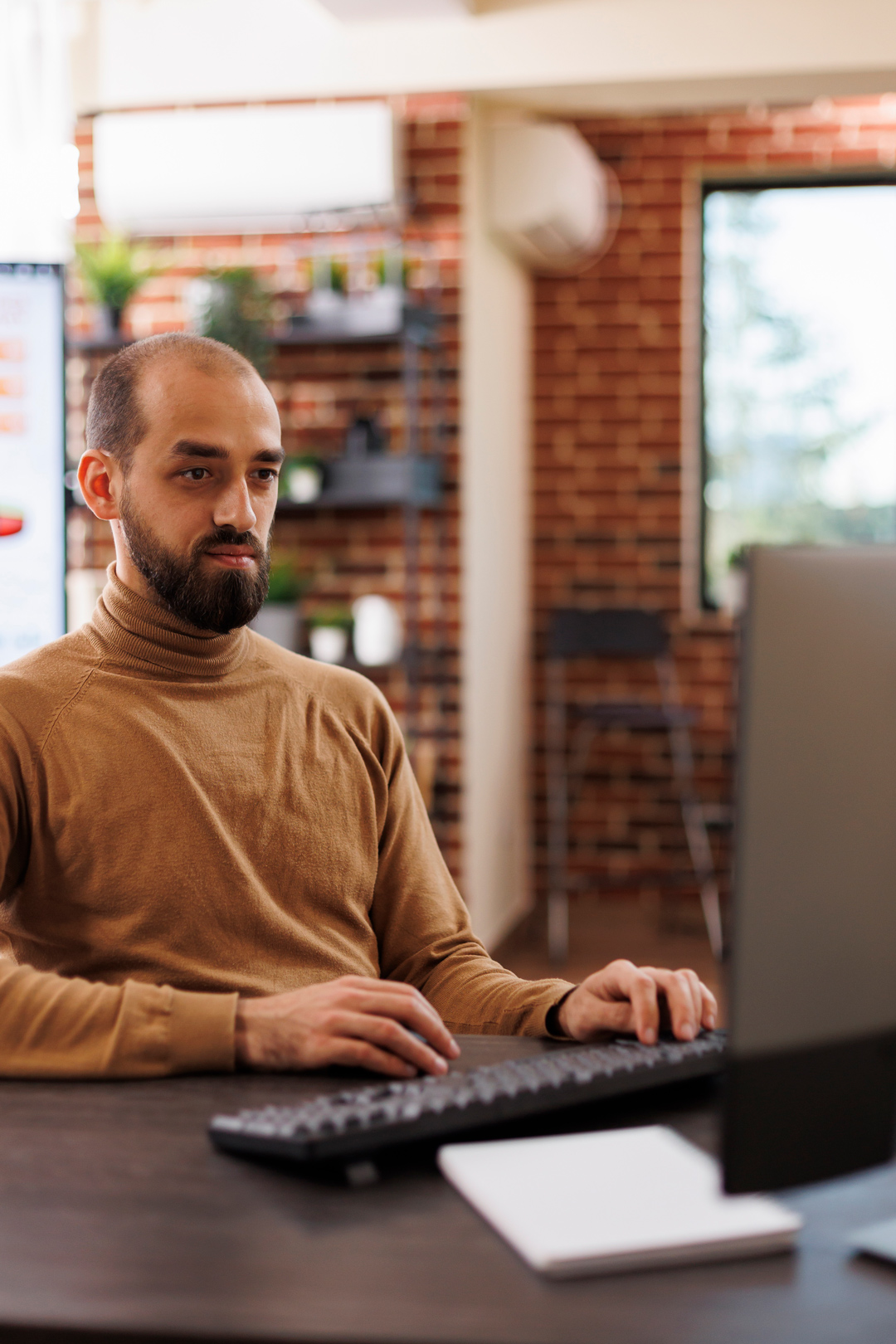 Man in a brown sweater working on a computer in a modern office setting, focused on his screen.