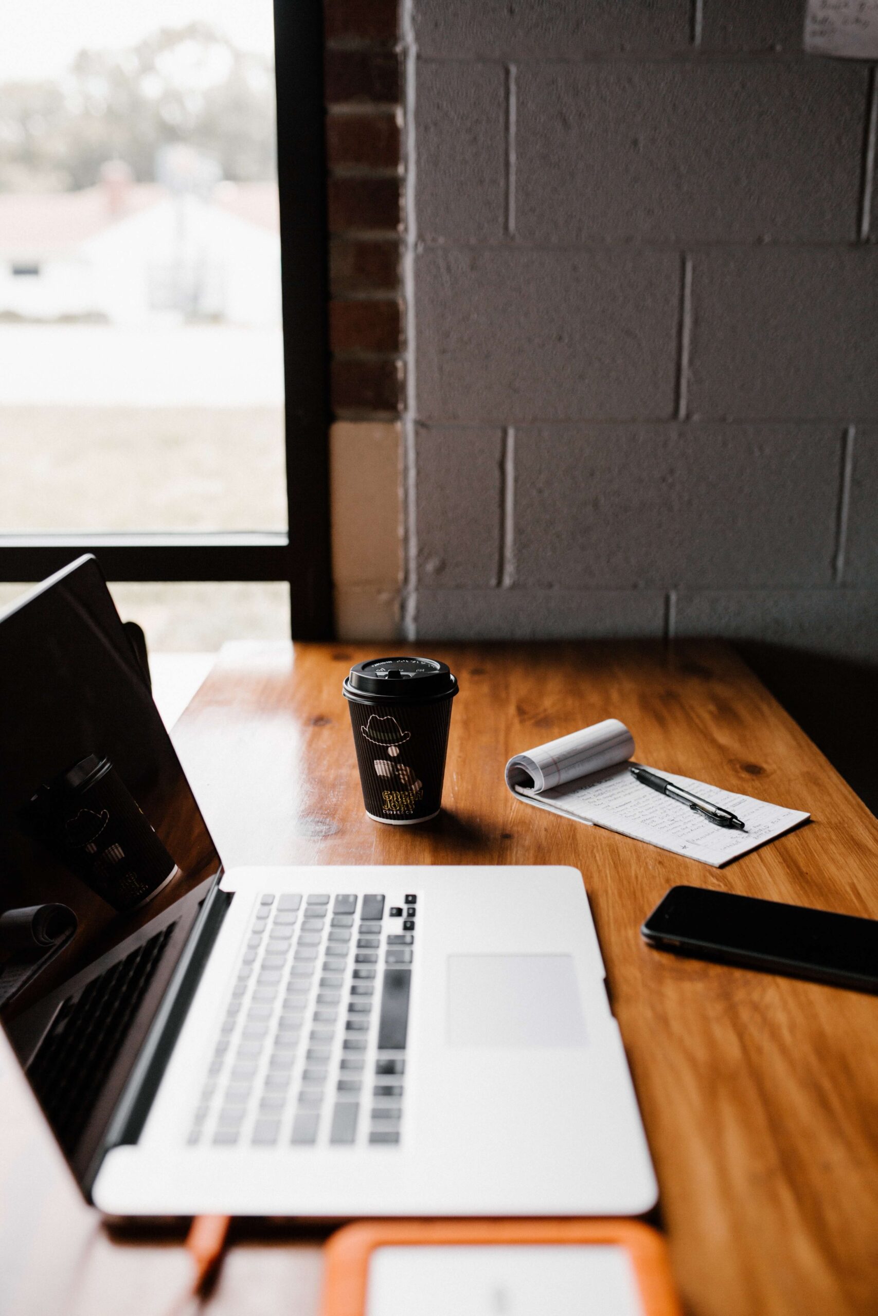 Cozy workspace with laptop, coffee cup, notebook, and pen near a window, perfect for productivity and creativity.