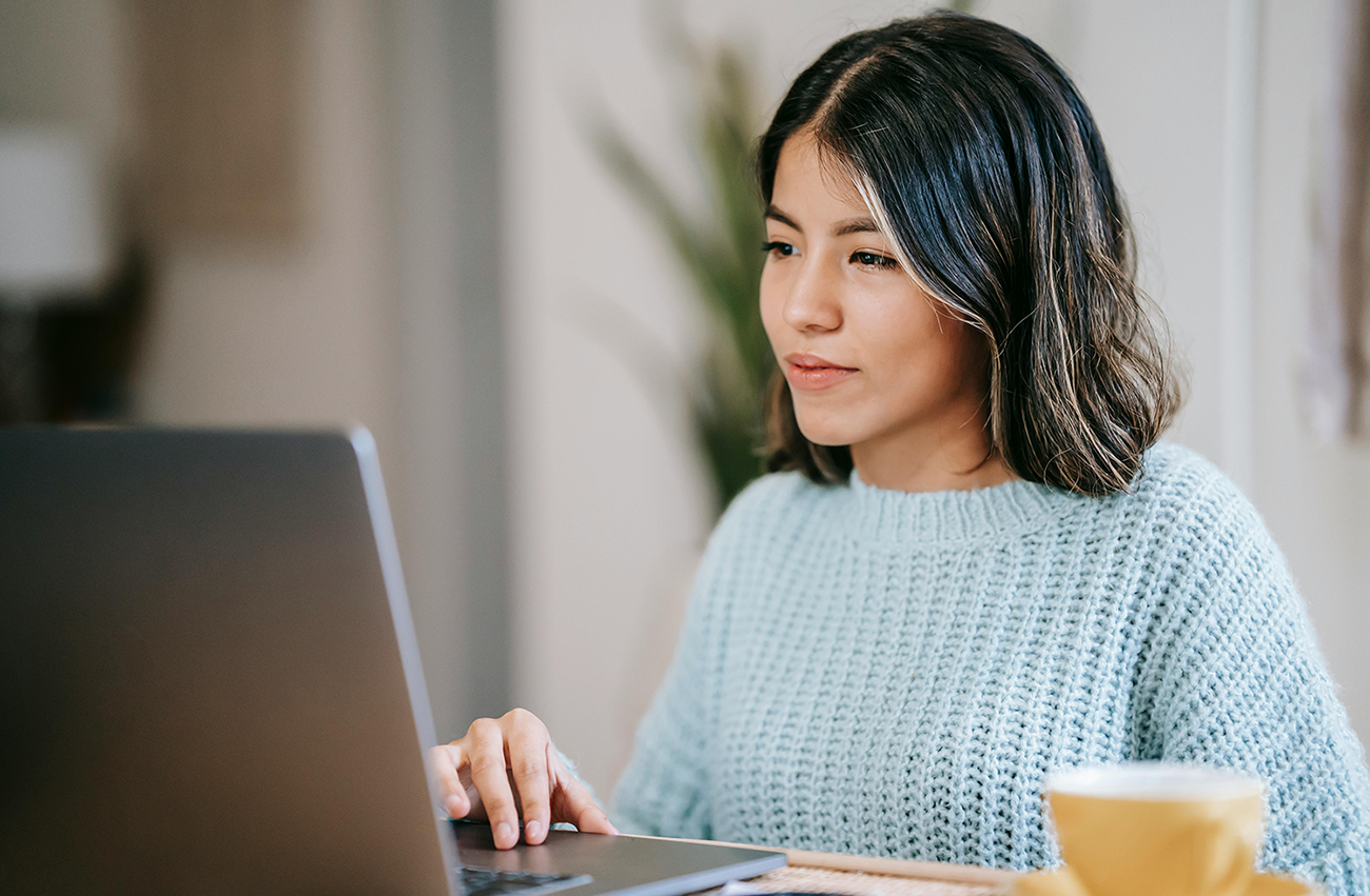 Woman in blue sweater working on a laptop at a cozy home setting with coffee cup nearby.