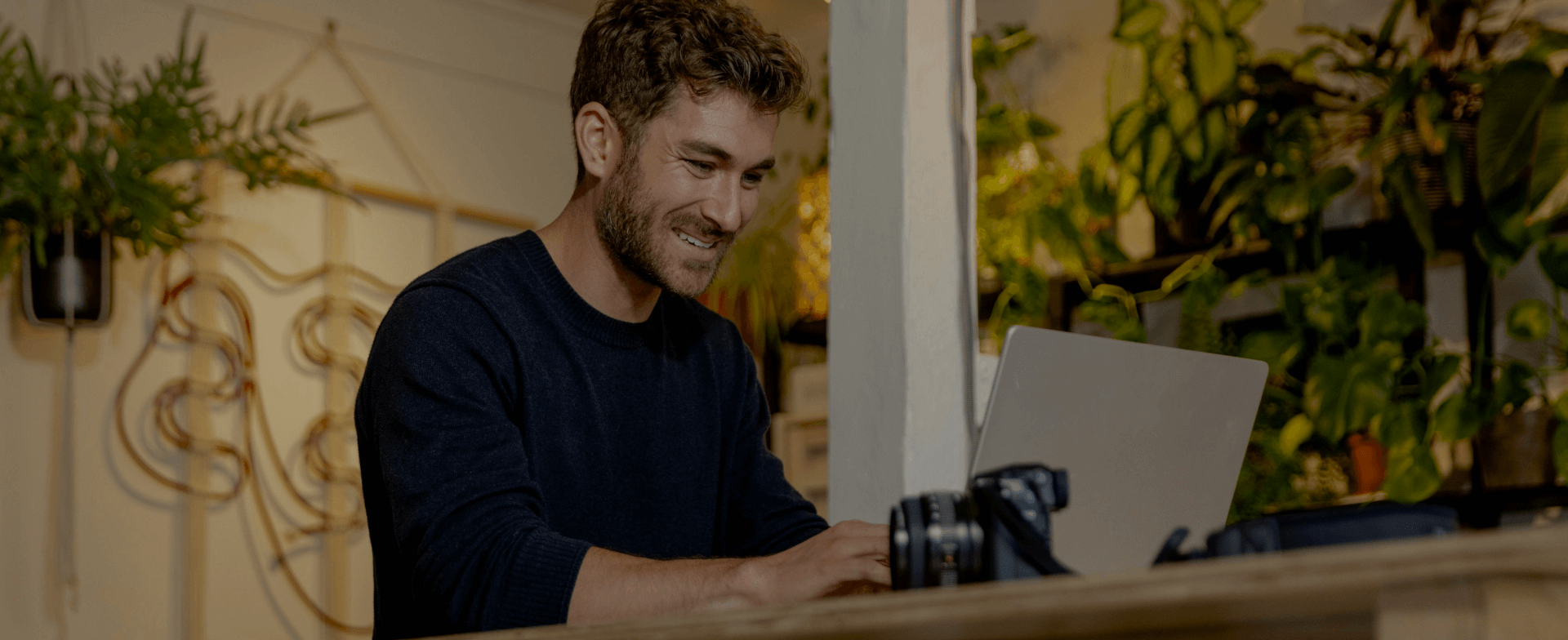 Smiling man working on laptop in home office with plants and camera nearby.