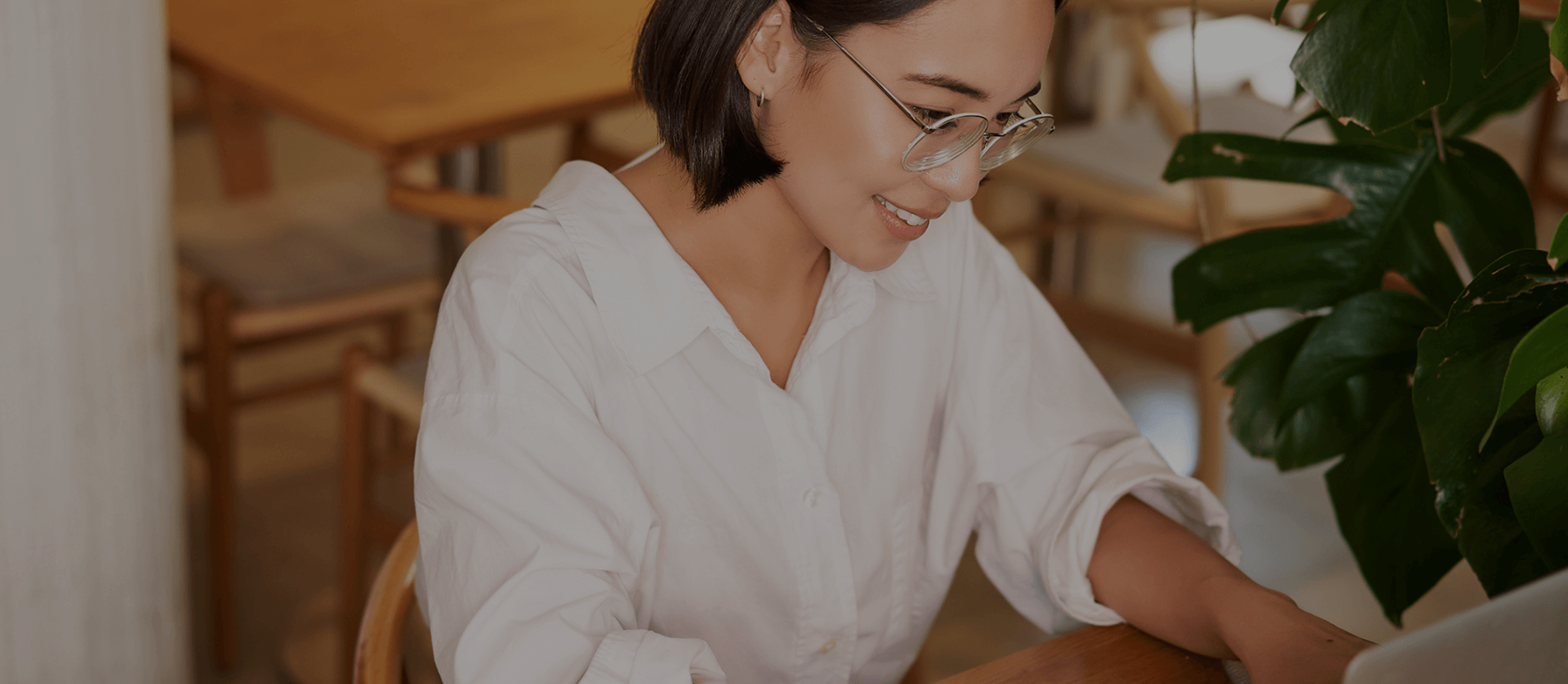 Woman in glasses working on a laptop in a cozy café setting with greenery.